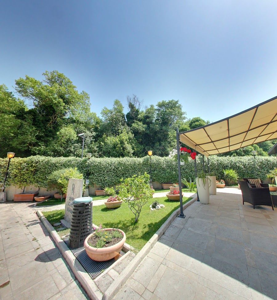 Patio esterno con piante in vaso, prato verde, alberi e ombrellone.