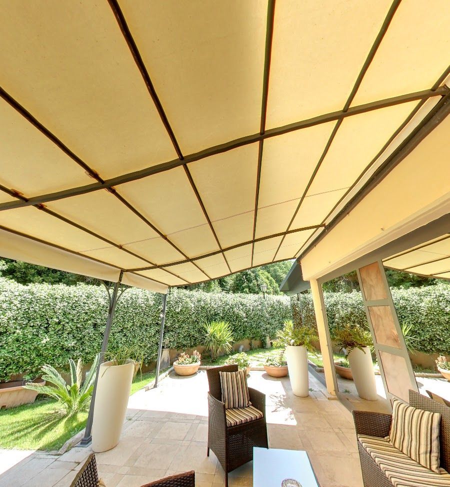 Patio con tenda da sole color crema sopra l'area salotto, vegetazione lussureggiante sullo sfondo.