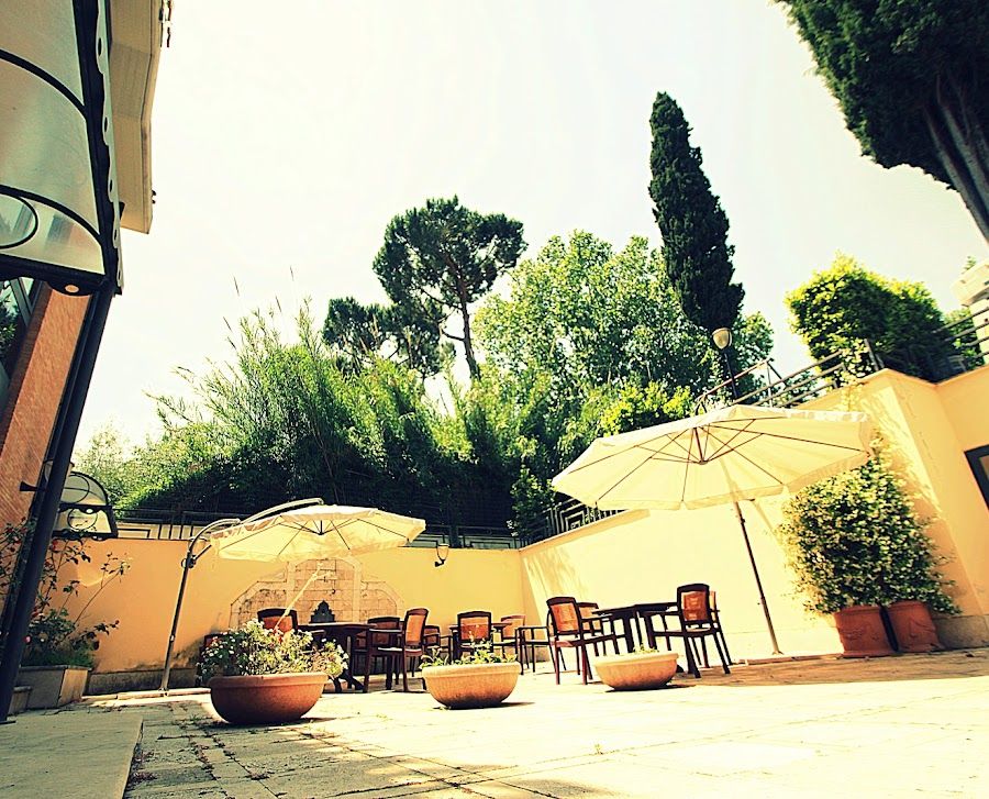 Patio esterno con tavoli, ombrelloni e piante in vaso, contro un muro con alberi sullo sfondo.