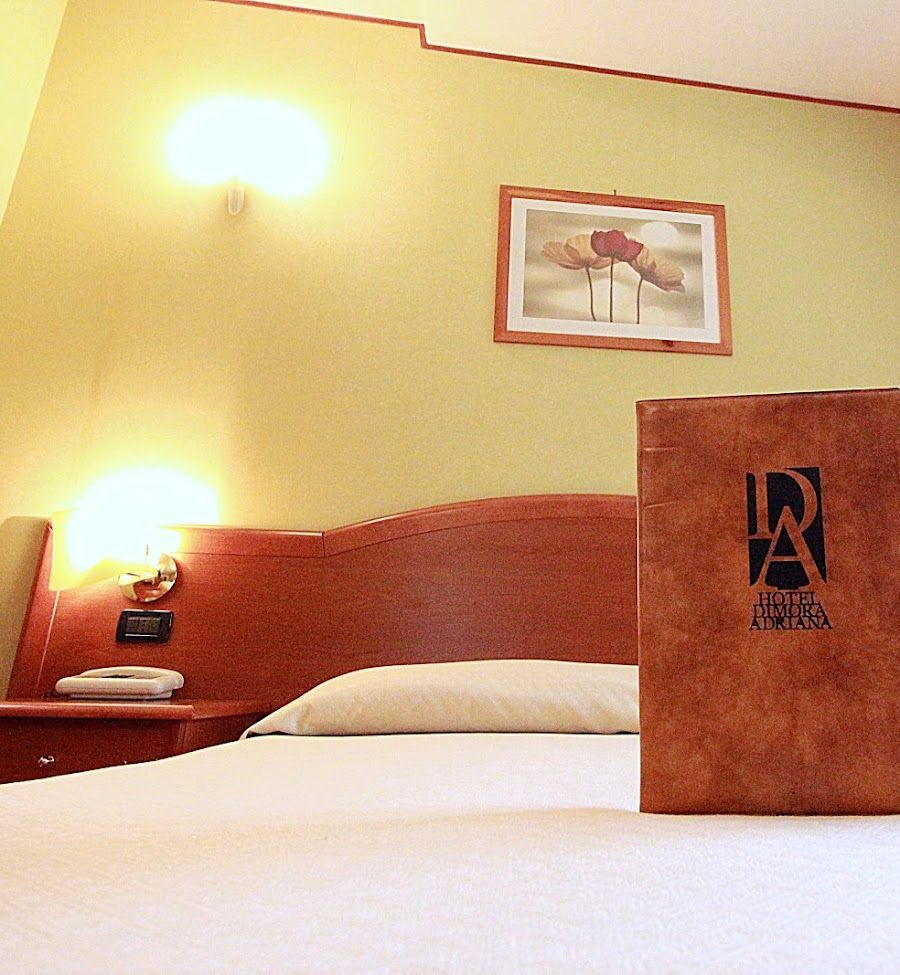 Interno di una camera d'albergo con letto, comodino e menù. Pareti verde chiaro, testiera in legno e luci soffuse.
