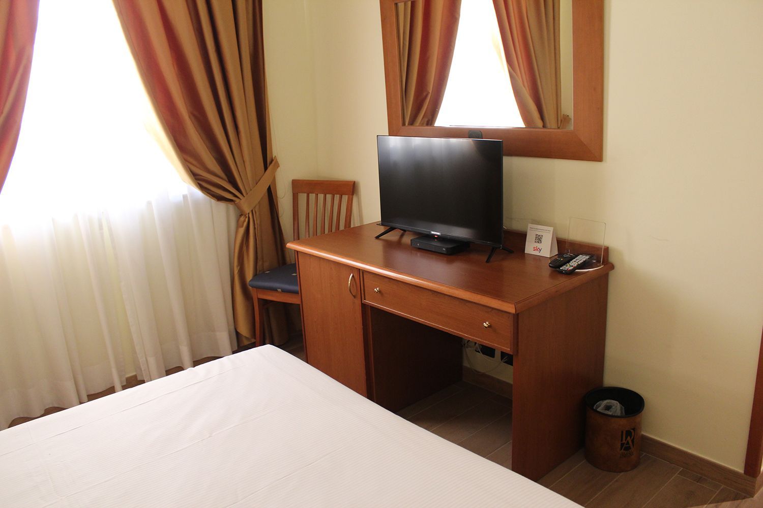Camera da letto con letto, scrivania, TV, finestra e sedia. Scrivania marrone, pareti beige e tende marroni.