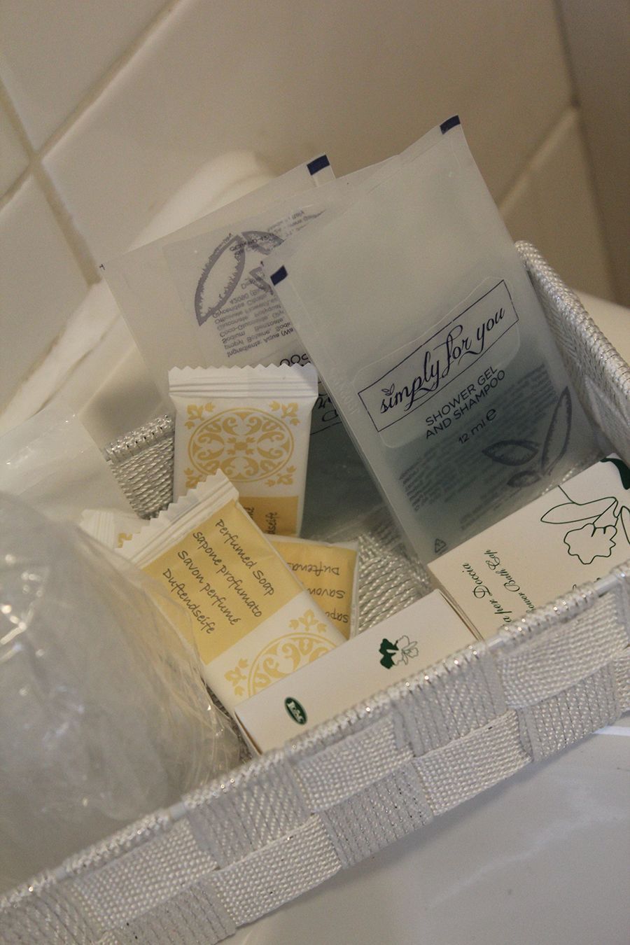 Un cestino contenente articoli da toeletta dell'hotel, tra cui bustine di sapone e shampoo, è appoggiato su una superficie bianca.