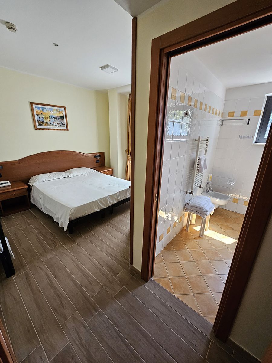 Camera d'albergo con letto e bagno visibili attraverso la porta. Pareti chiare, pavimento in legno e piastrelle color cuoio nel bagno.