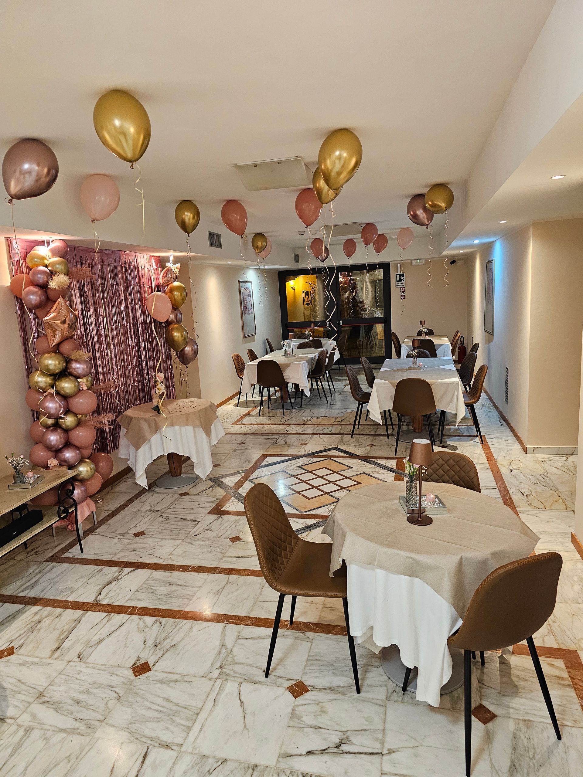 Ristorante decorato con palloncini dorati e rosa, tavoli apparecchiati, pavimento in marmo, decorazioni per la festa.