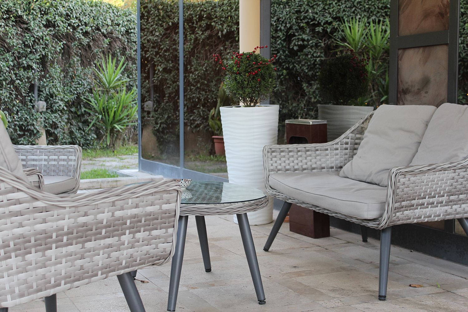 Patio con mobili in vimini, tavolo con piano in vetro e piante contro una parete verde.