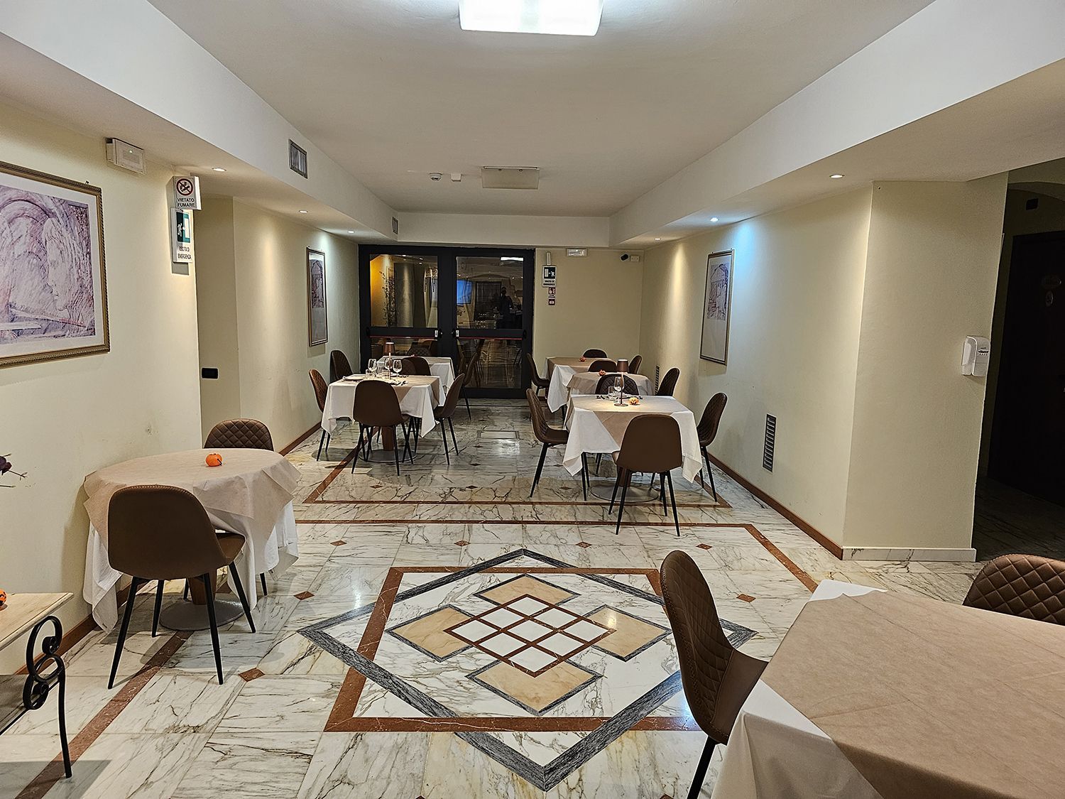 Interno di un ristorante con tavoli apparecchiati per cenare, pavimento in marmo, colori neutri.