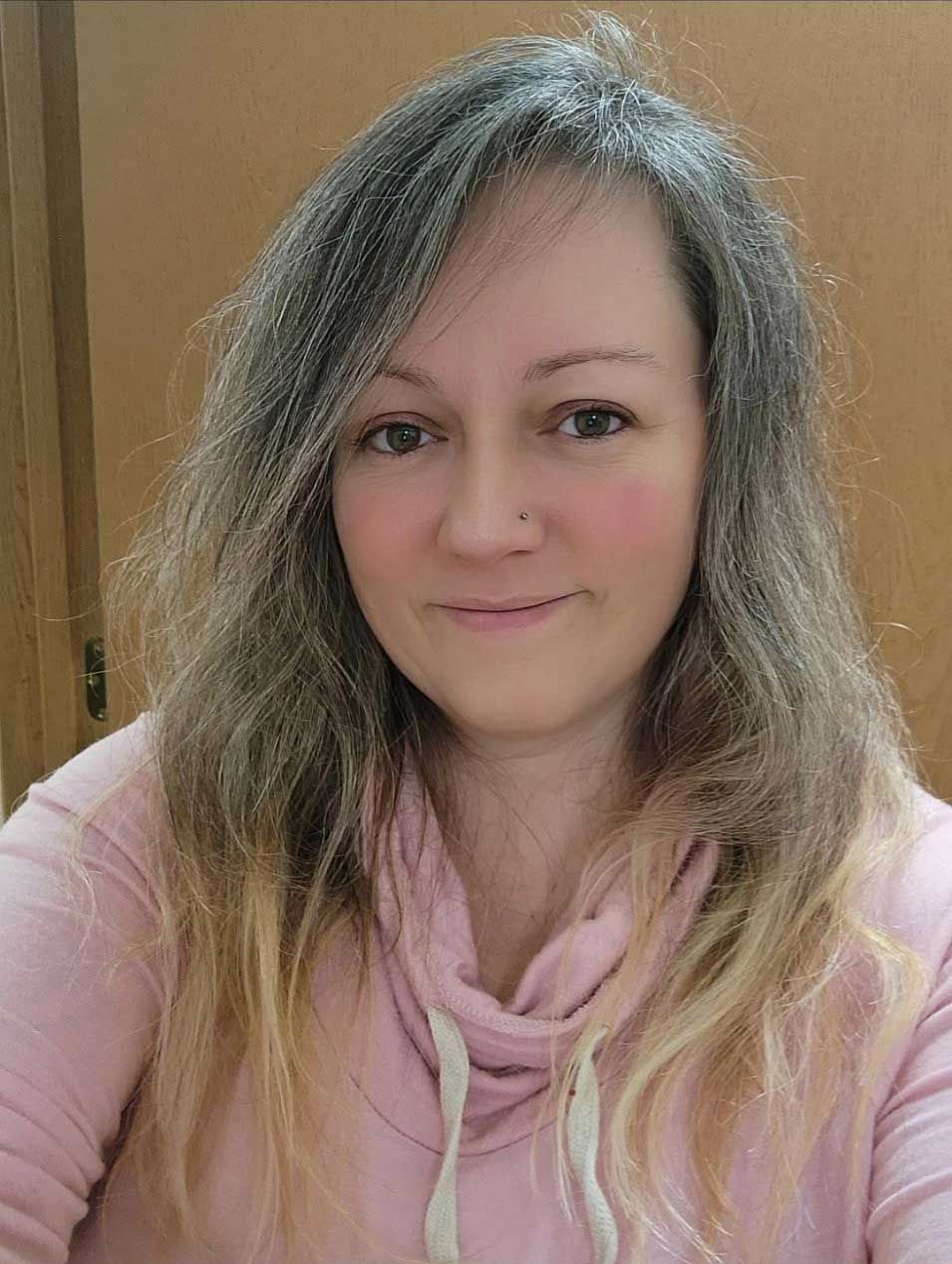 Person with long gray-blonde hair wearing a pink hoodie, smiling against a plain beige background