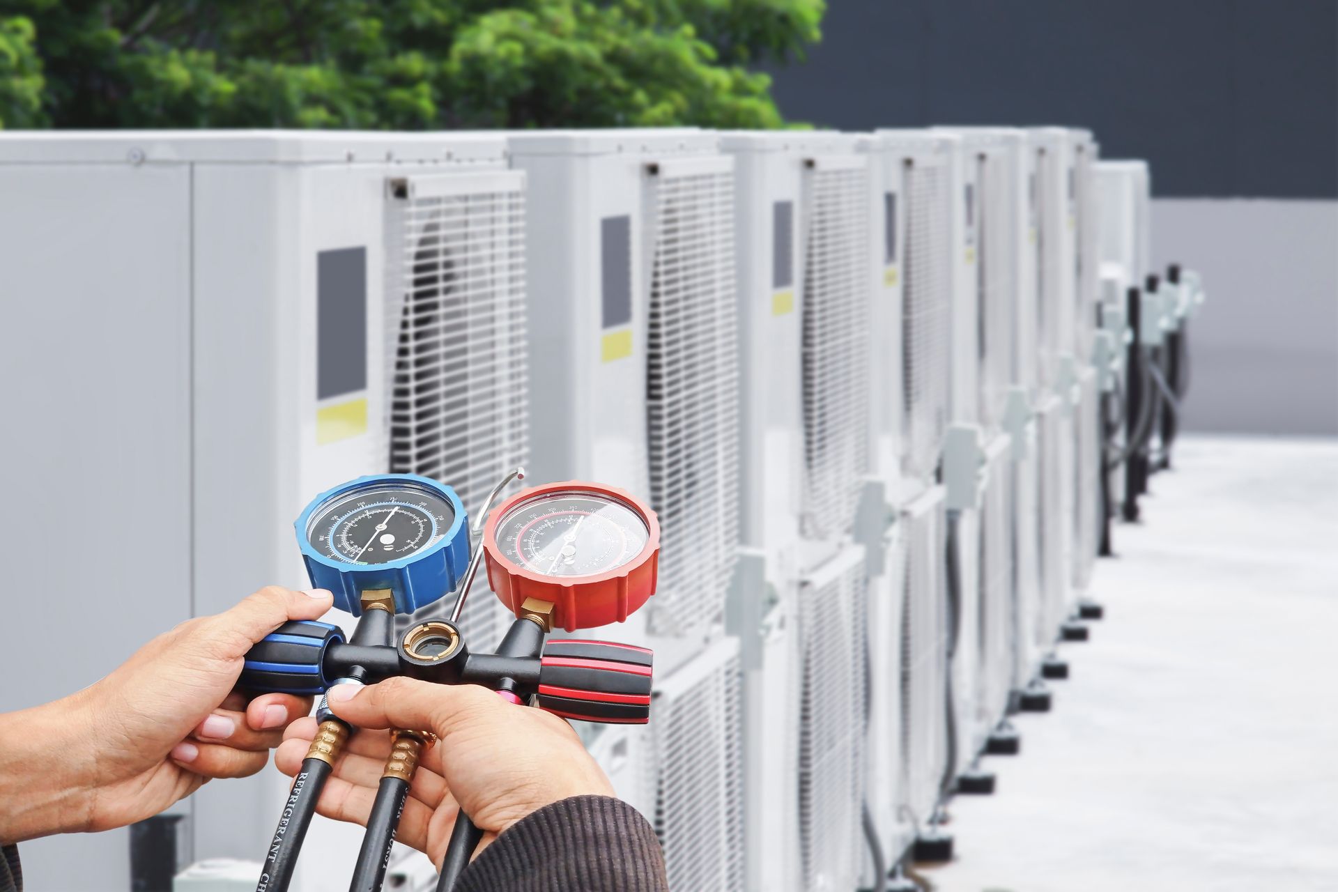 Technician holding gauges beside outdoor air conditioning units during HVAC maintenance