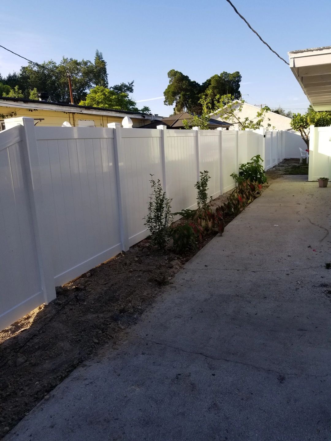 A white fence surrounds a concrete driveway in a backyard.