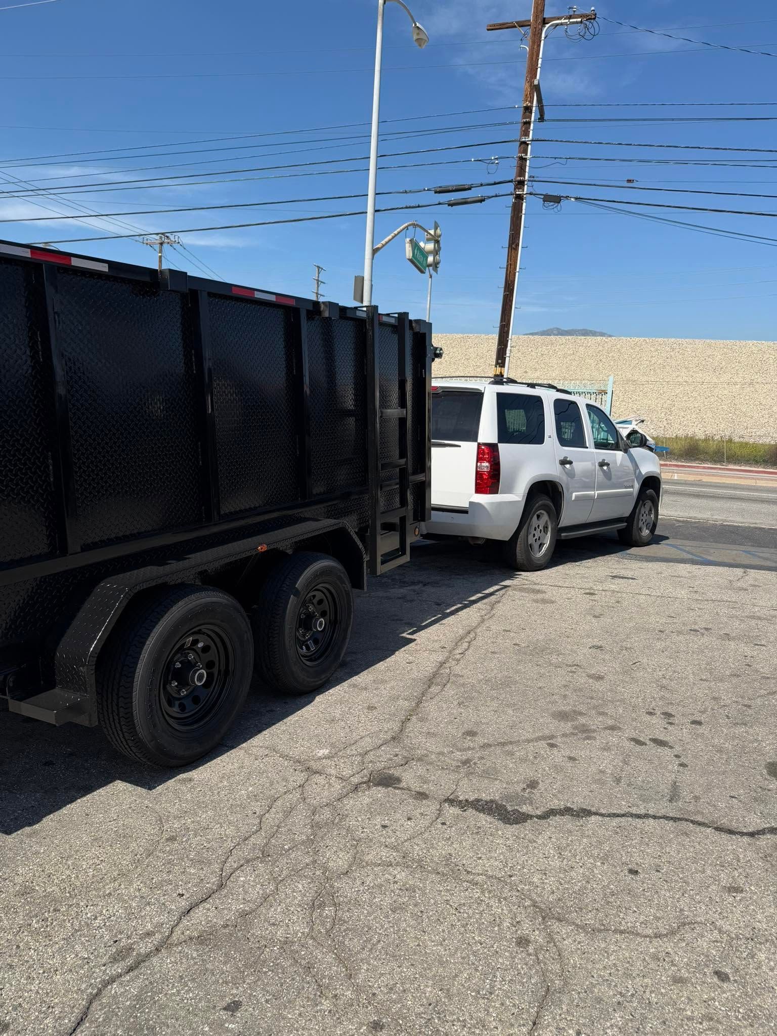 White SUV hitched to a black dump trailer on a gravel roadside under a blue sky.