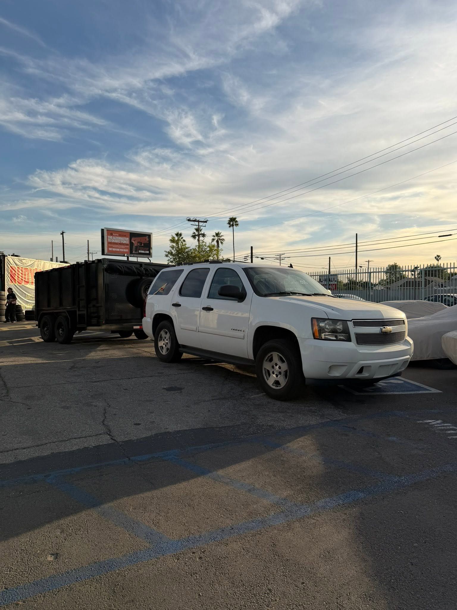 White SUV towing a black trailer in a parking lot at sunset under a cloudy sky