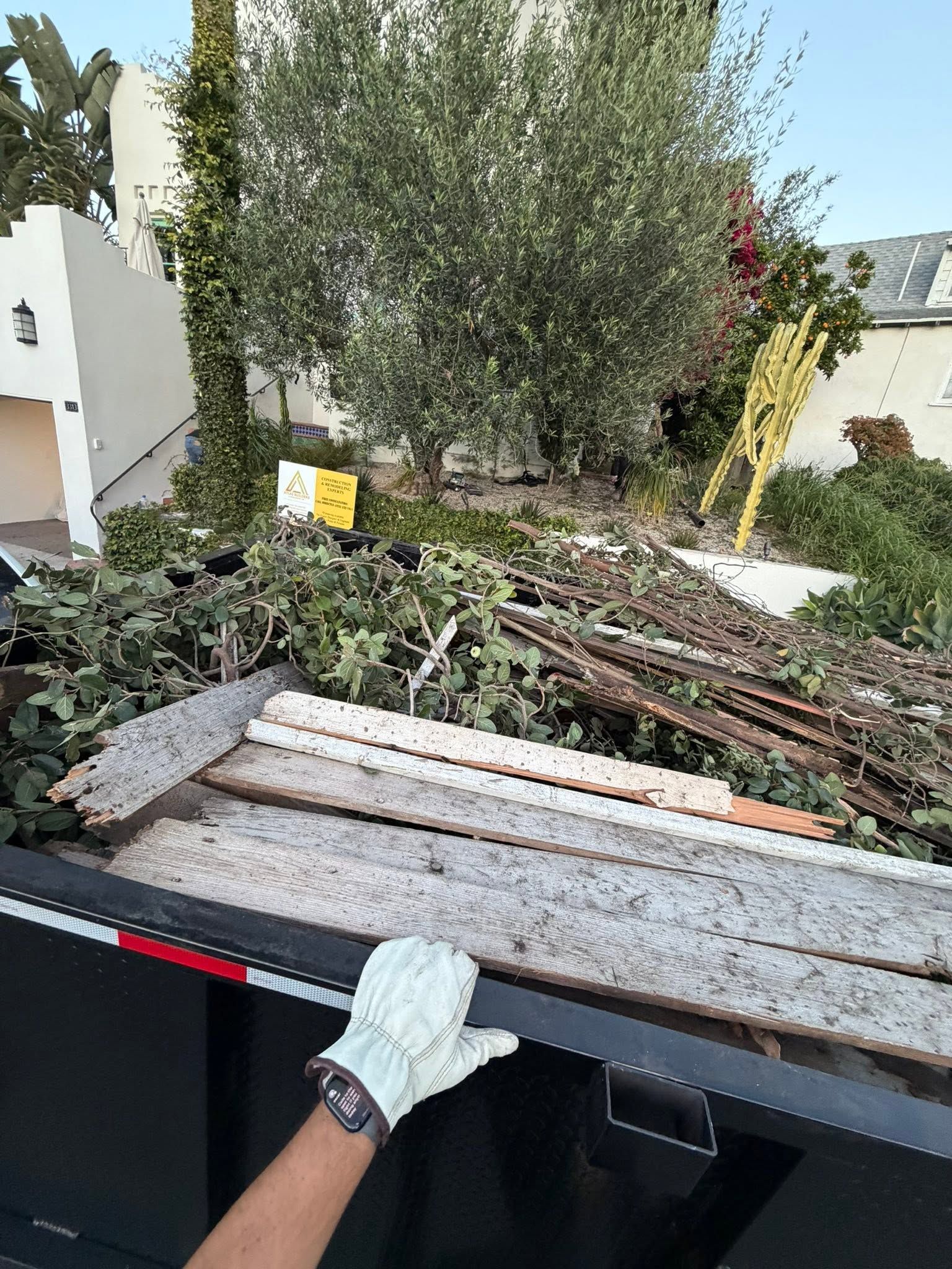 Gloved hand loading stacked wooden planks into a black truck bed in a garden with trees and debris