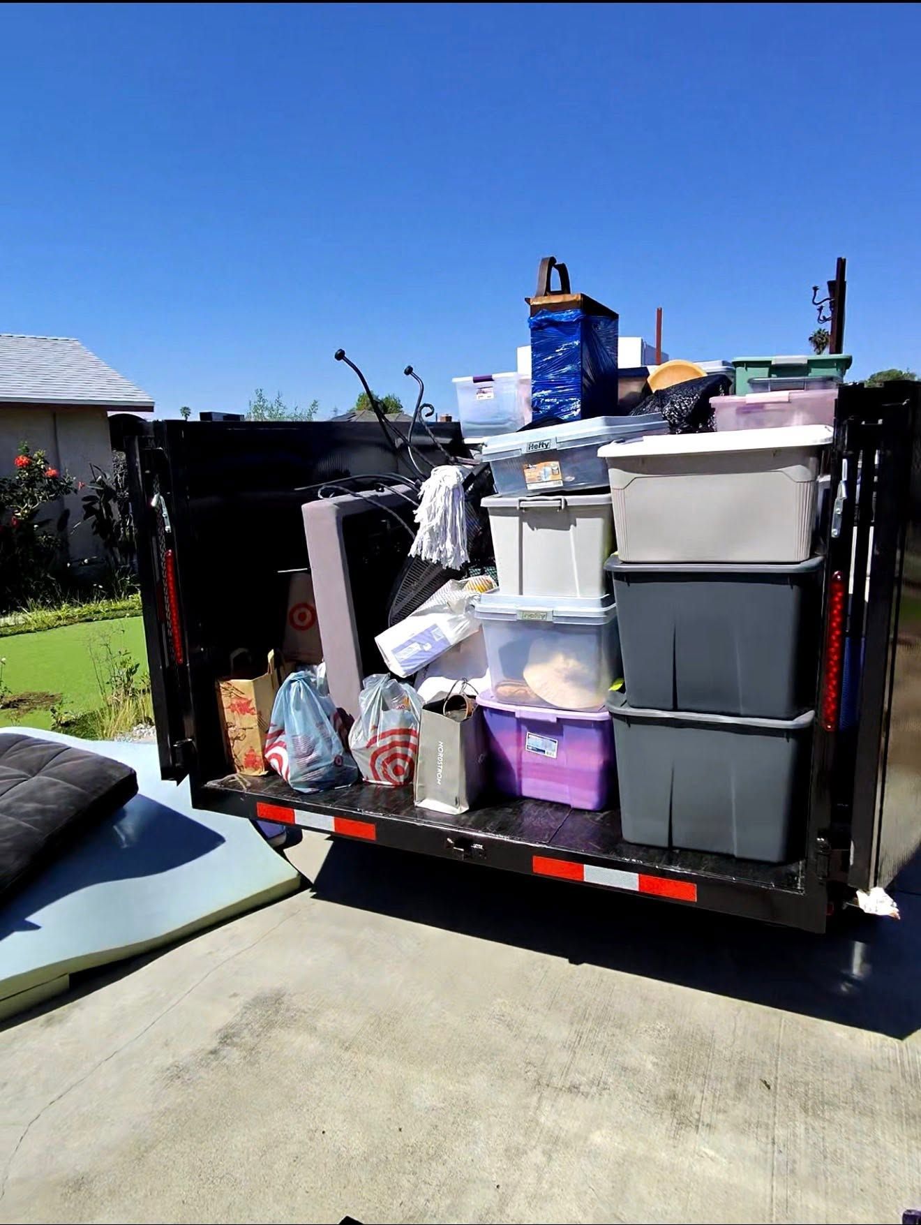 Stacked storage bins and household items loaded in the back of a black trailer on a sunny driveway.