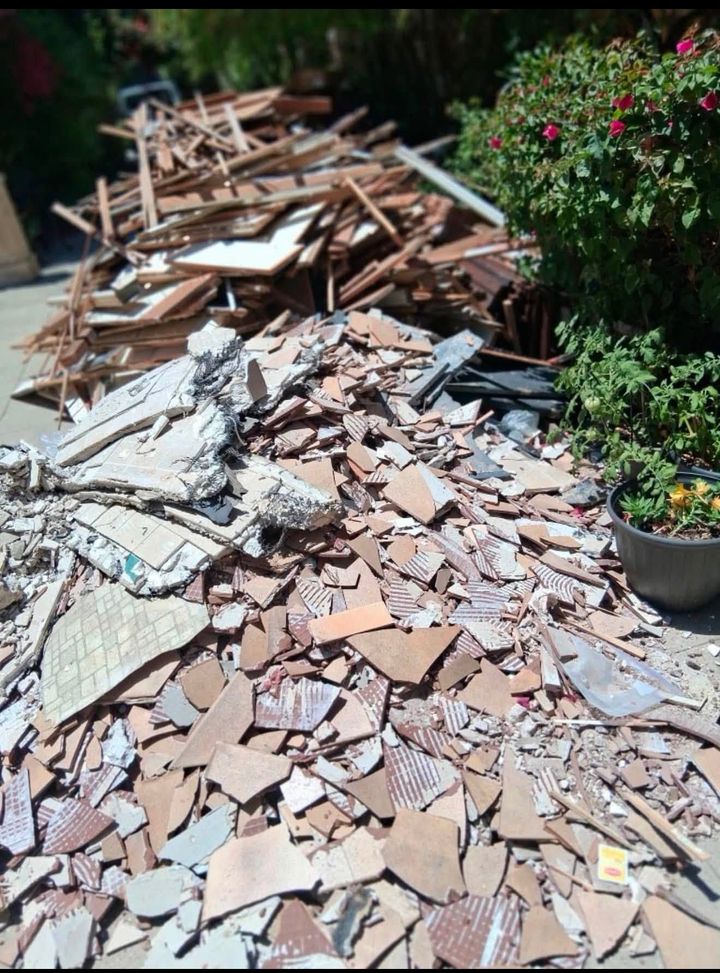 Pile of broken tiles and rubble scattered across a sunlit driveway beside green bushes.