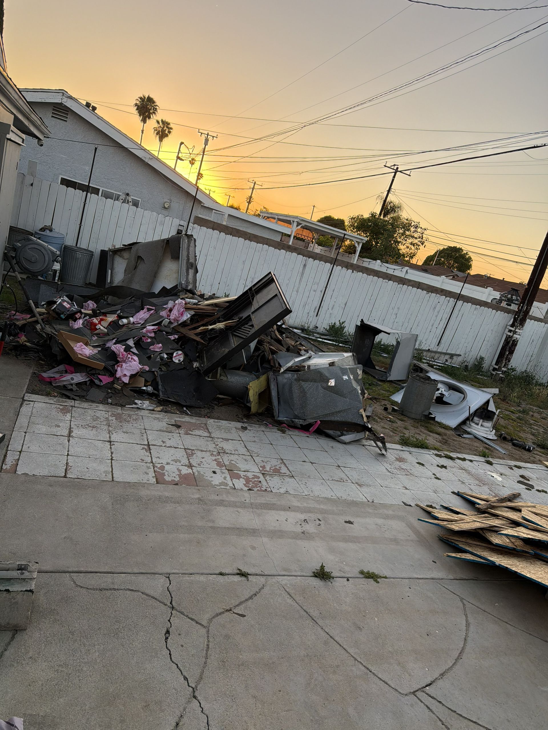 Debris piled beside a cracked concrete driveway at sunset, with a damaged wall and string lights overhead