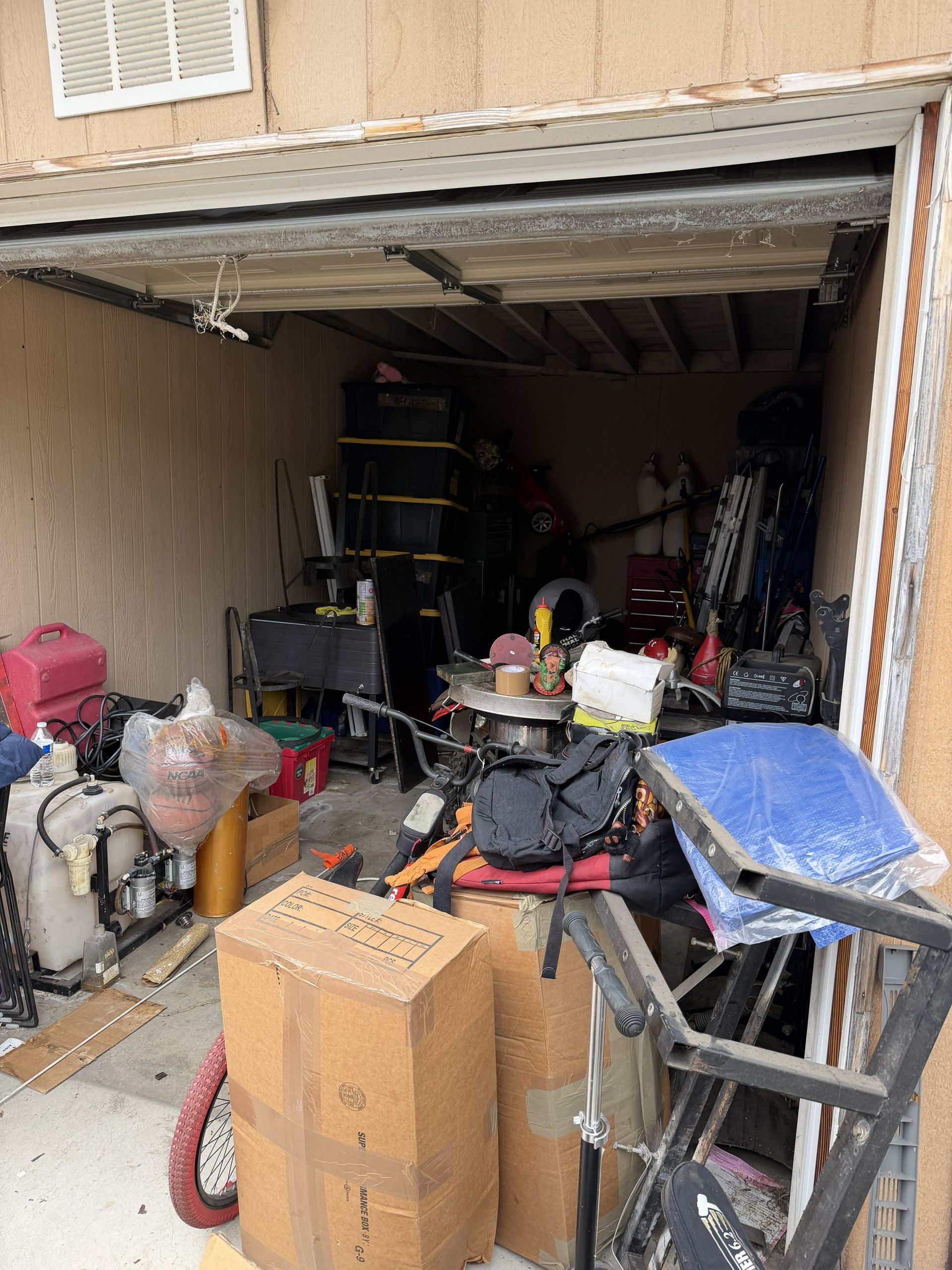 Cluttered open garage filled with boxes, tools, and household items, with a partially raised door.