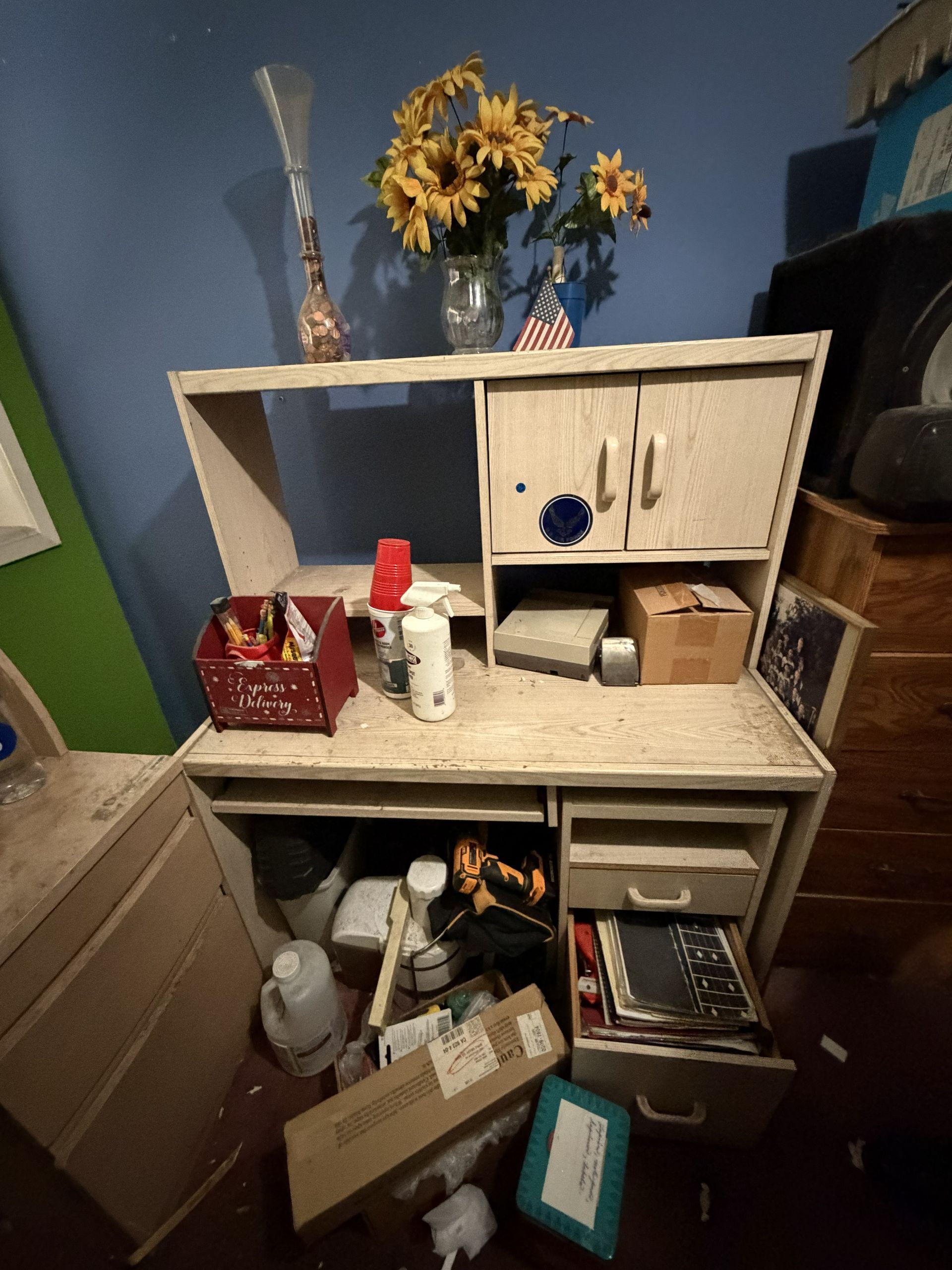 Cluttered kitchen hutch with scattered dishes, boxes, and small items; sunflowers on top against a blue wall