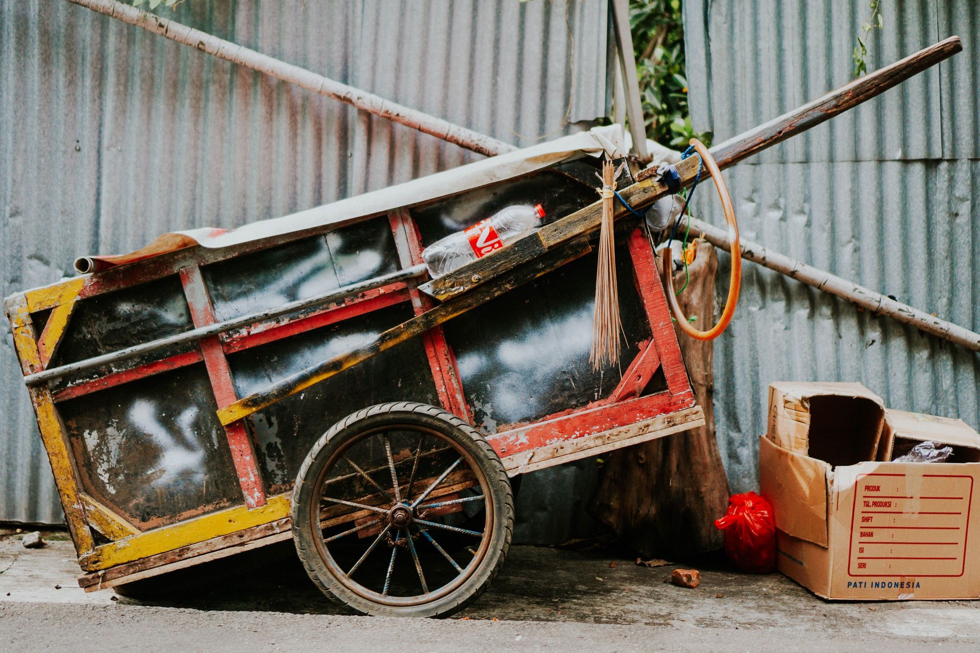 A damaged red cart leaning against a corrugated metal wall, with boxes and debris beside it.