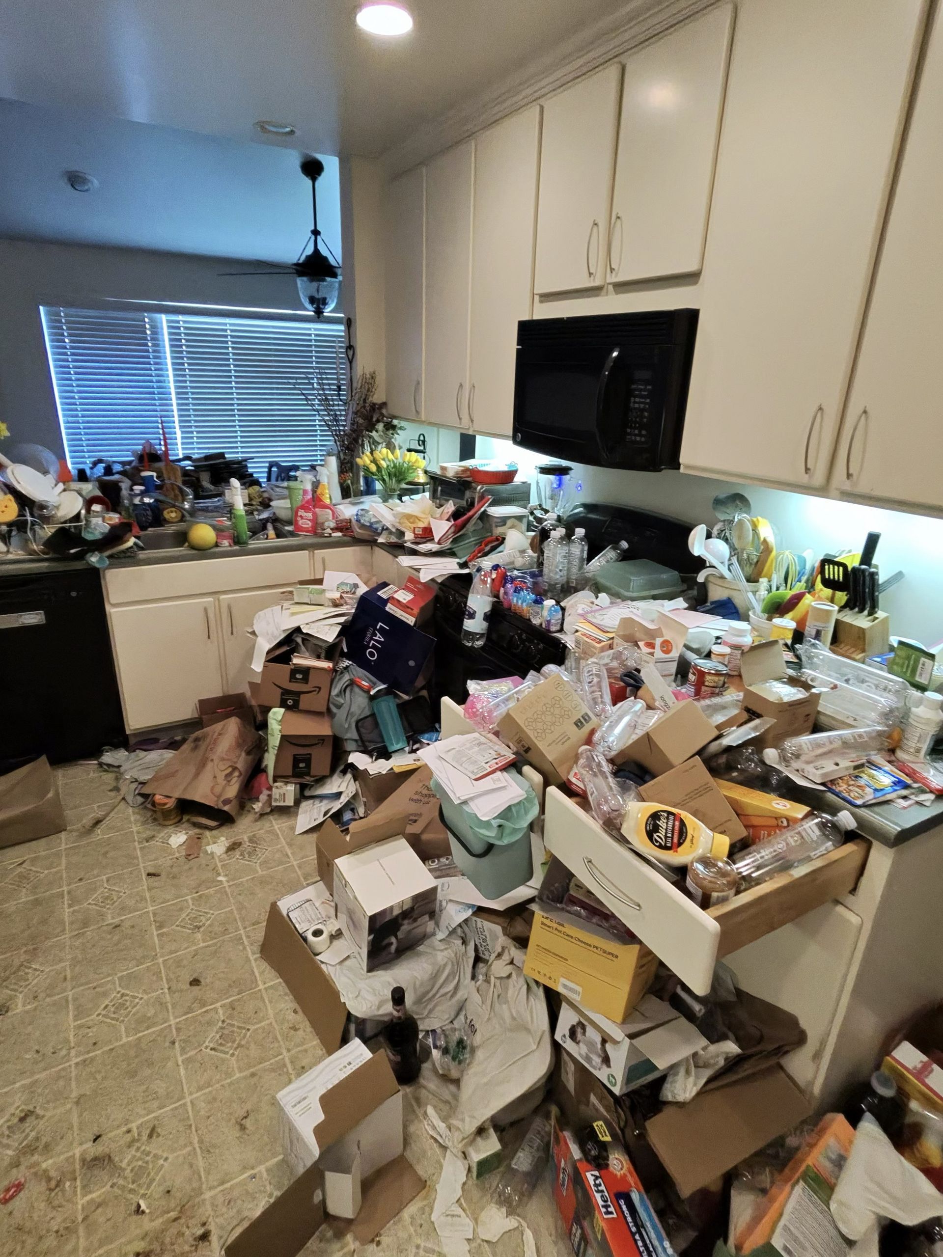 Cluttered kitchen with boxes and supplies piled on the floor and counters, sink and cabinets visible.