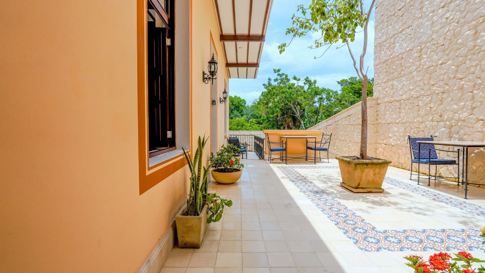 Patio exterior soleado con paredes beige, plantas en macetas y un camino de baldosas que conduce a una zona de estar