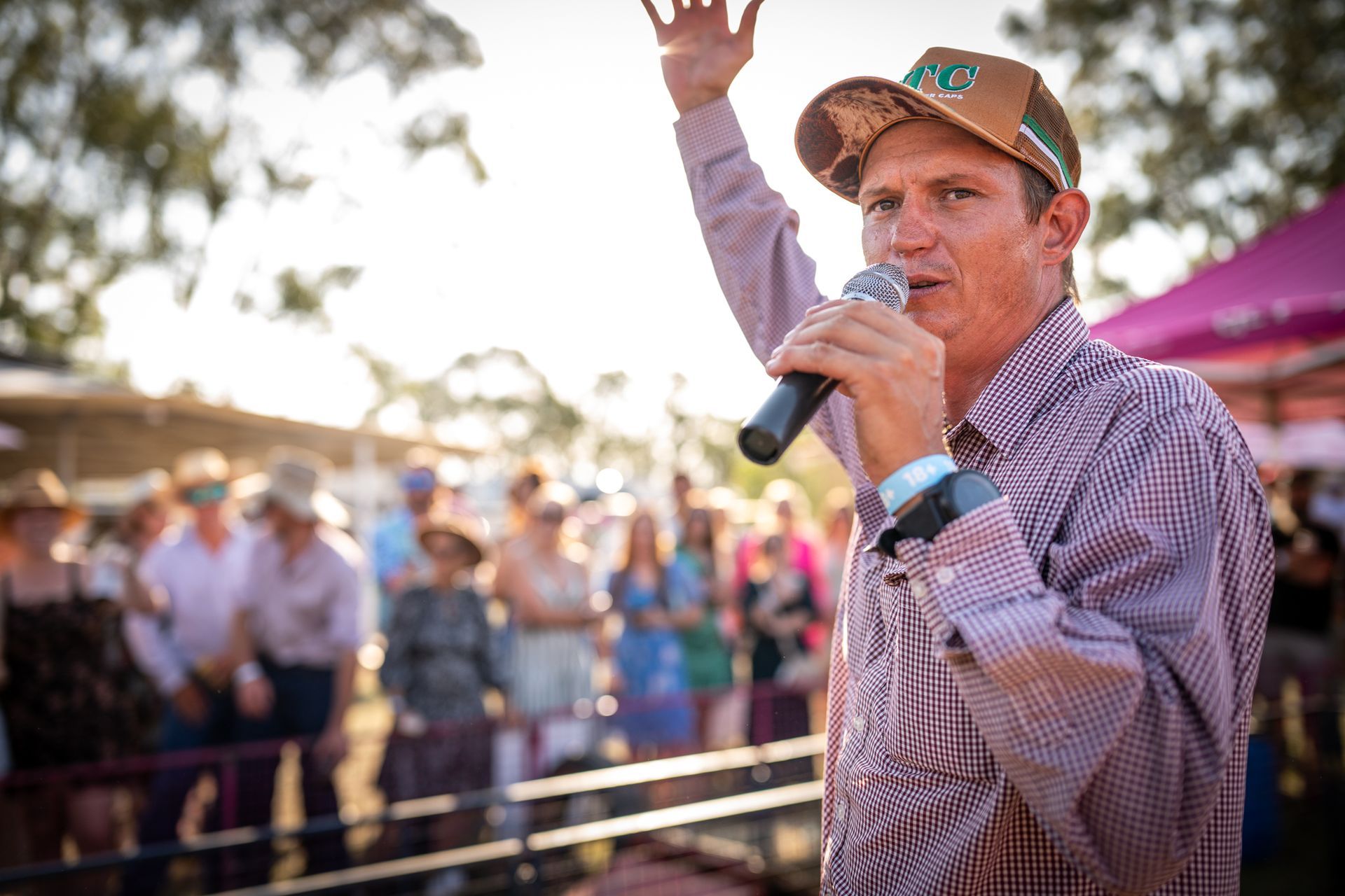 Man speaking into a microphone at an outdoor event, raising one hand to a crowd behind him.
