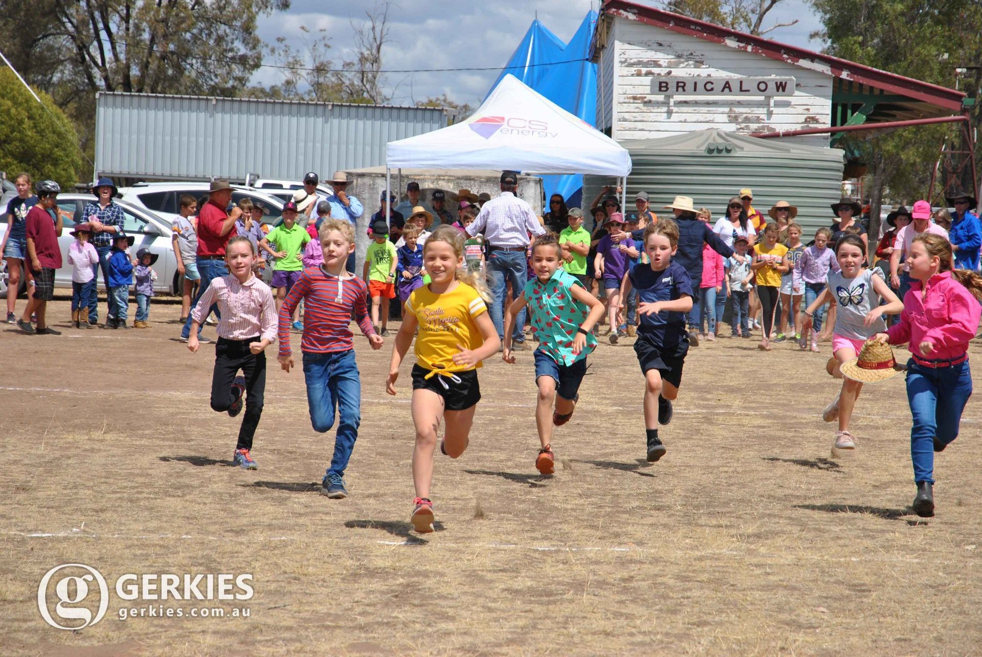 Children racing barefoot in colorful shirts at an outdoor field event with spectators and tents behind them