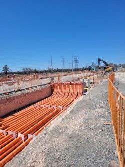 Construction site with orange cables laid in trenches beside a fenced gravel path and excavator in the distance