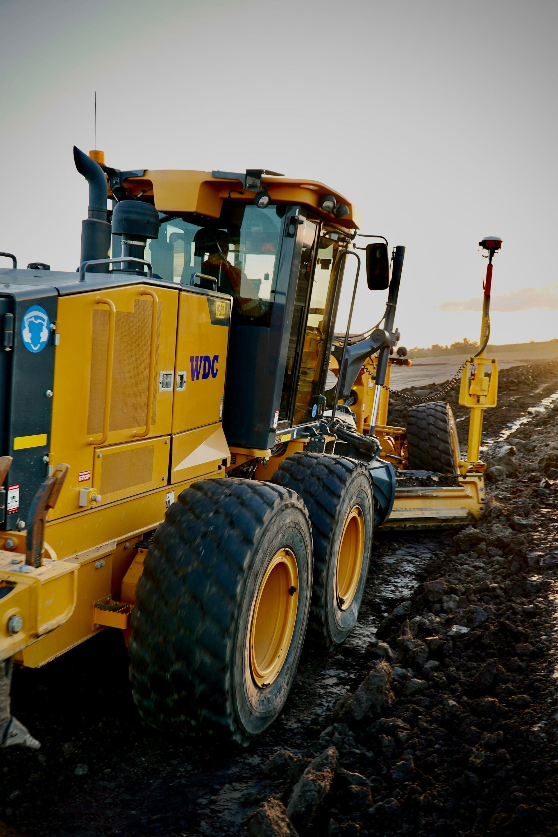 Yellow road grader on a muddy construction site under an overcast sky