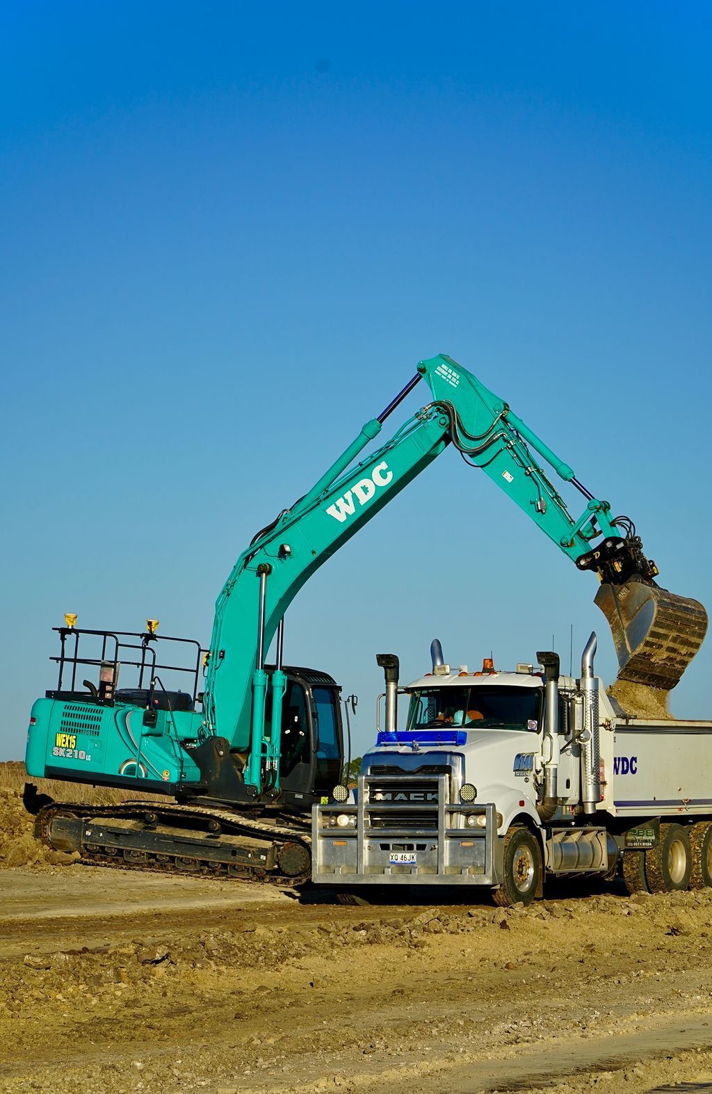 Green excavator loading dirt into a white dump truck on a construction site under a clear blue sky