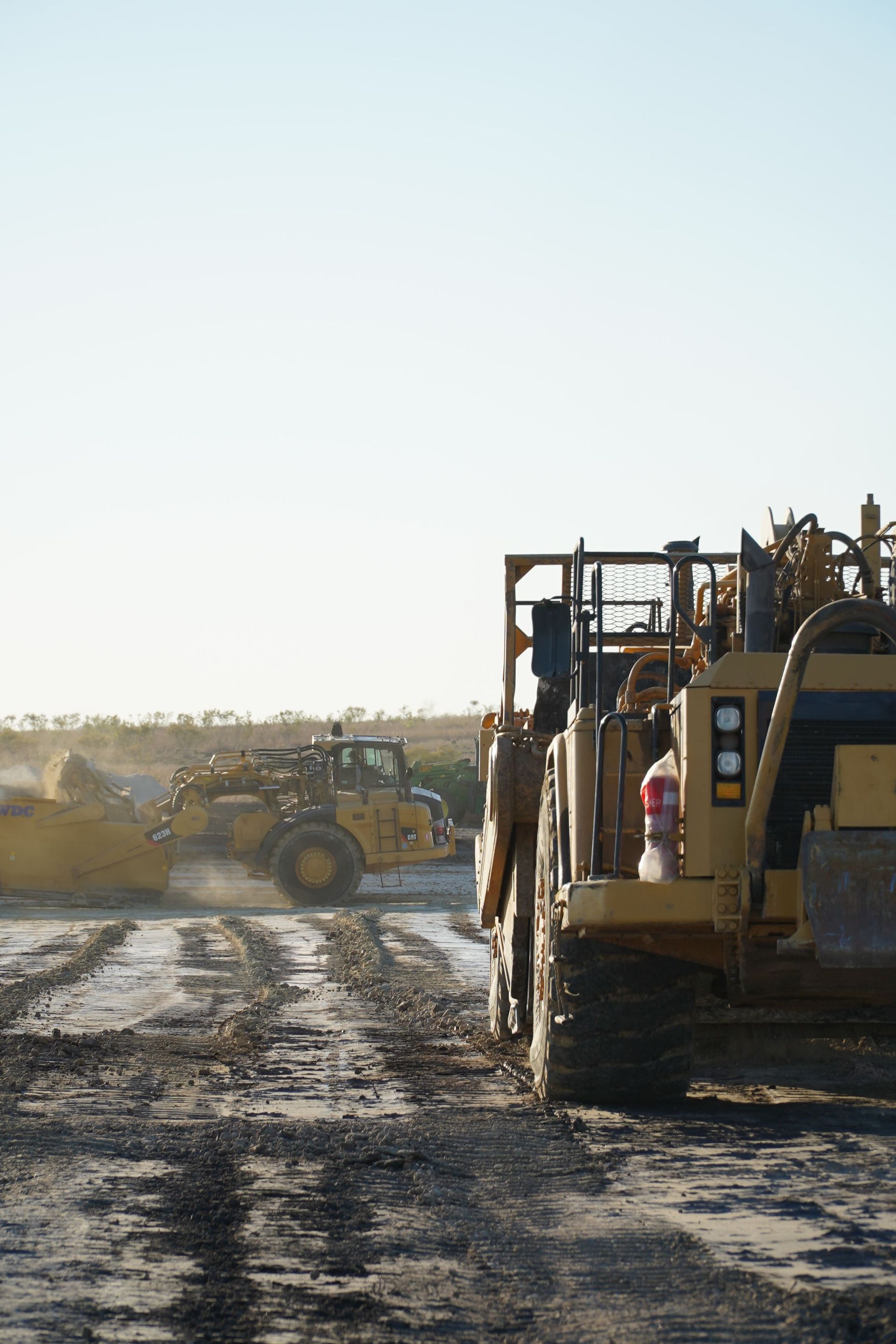 Construction vehicles on a muddy dirt road at a worksite under a bright sky