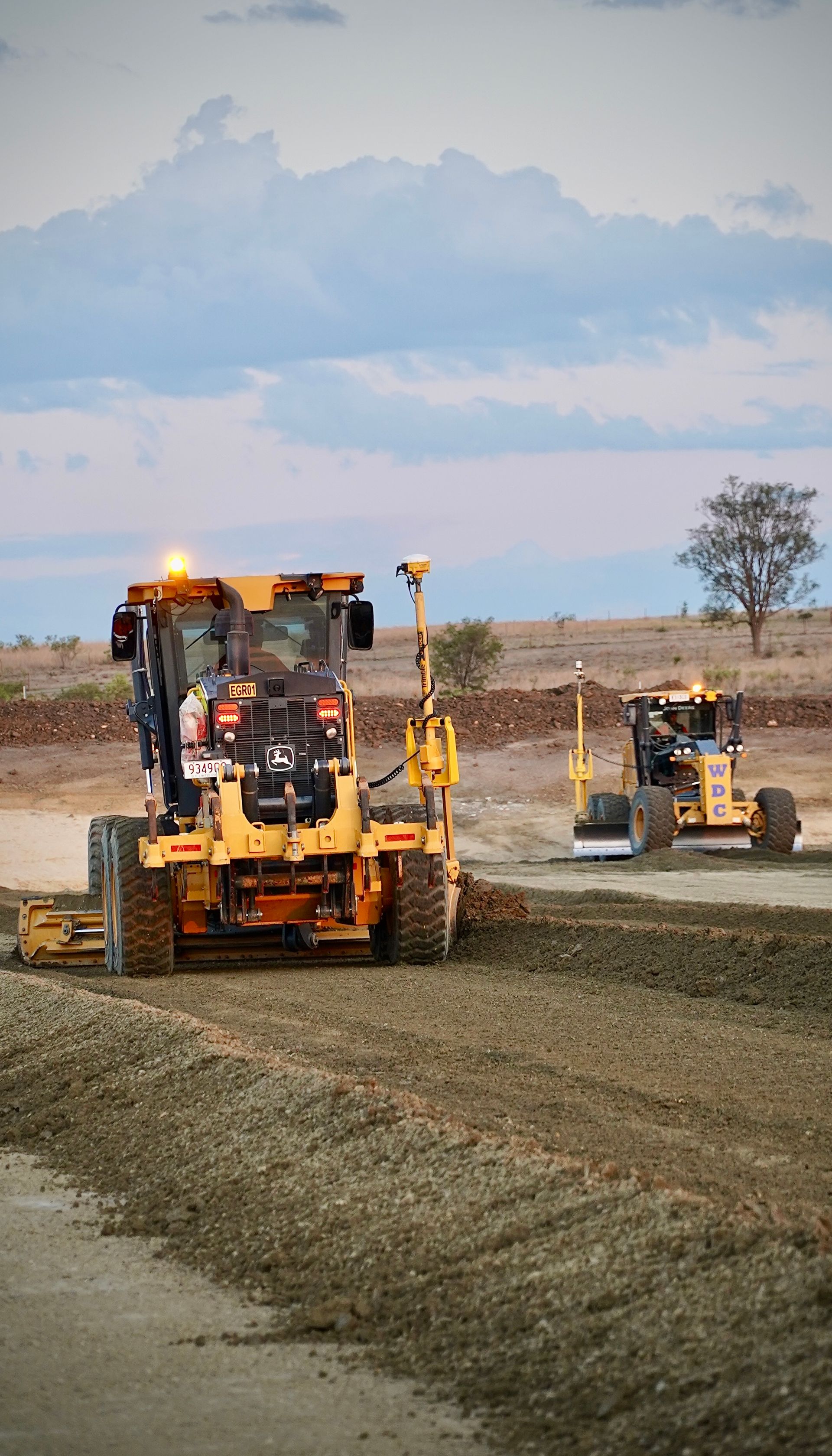Two road graders smoothing gravel on a dirt road under a cloudy sky