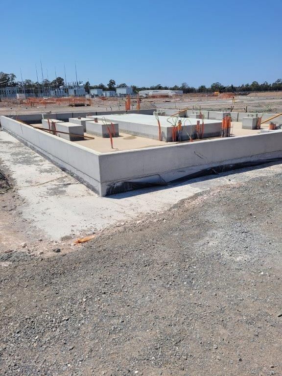 Concrete foundation slab under construction on a gravel site, with exposed rebar and pipes in an open area.