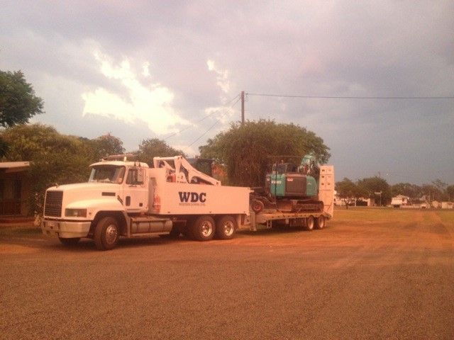 White WDC truck towing a green machine on a flatbed through a dusty dirt lot at sunset