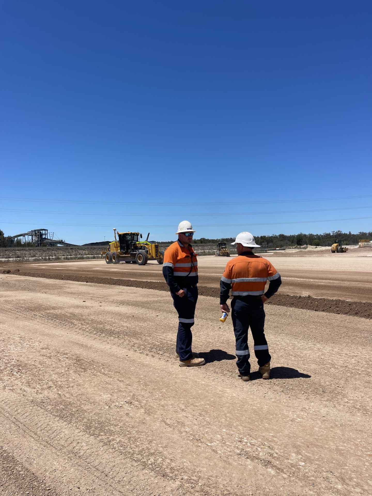 Two construction workers in hard hats standing on a gravel road at a roadwork site under a clear blue sky