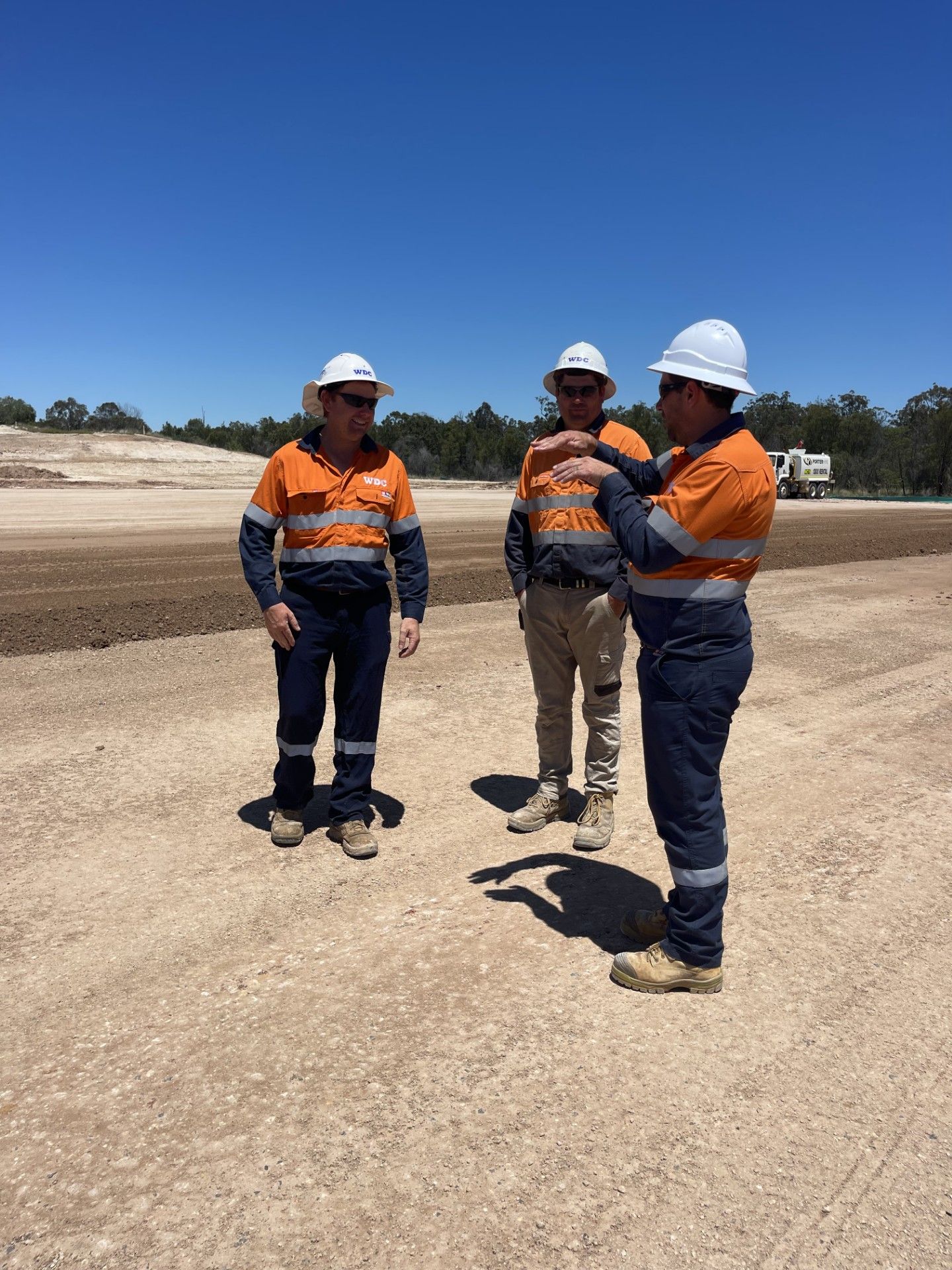 Three construction workers in hard hats and orange shirts standing on a sandy site under a clear blue sky