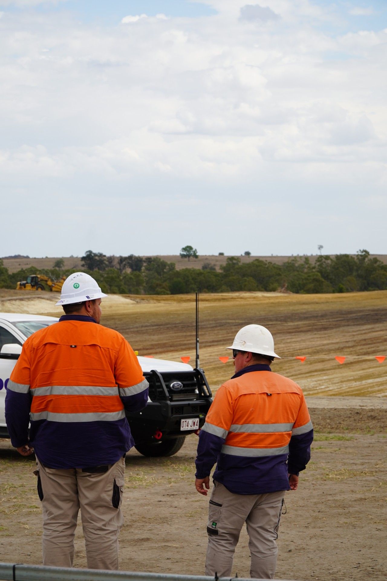 Two workers in orange safety jackets and white hard hats stand near a car on a construction site.