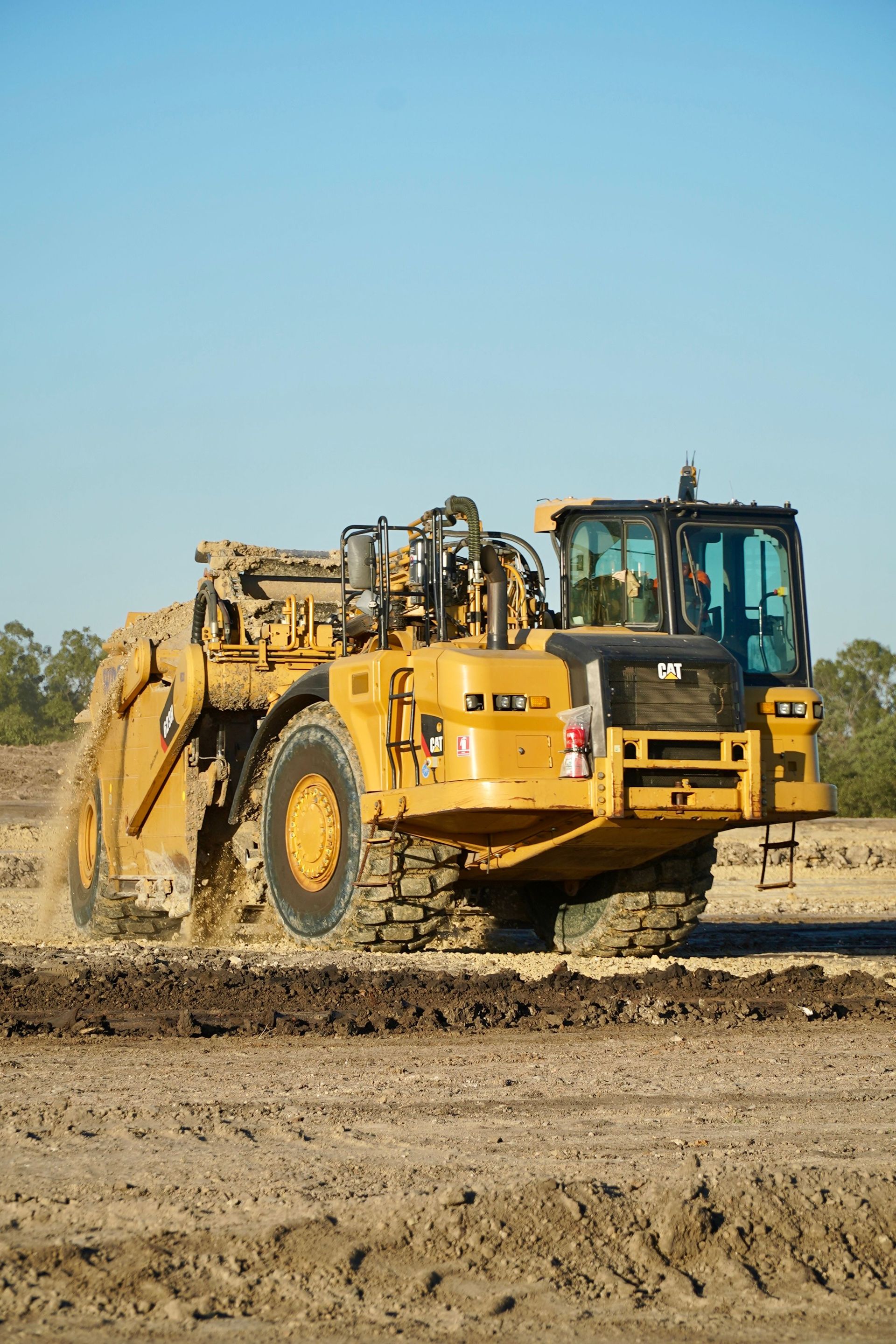 Yellow construction scraper moving soil on a dirt road under a clear blue sky
