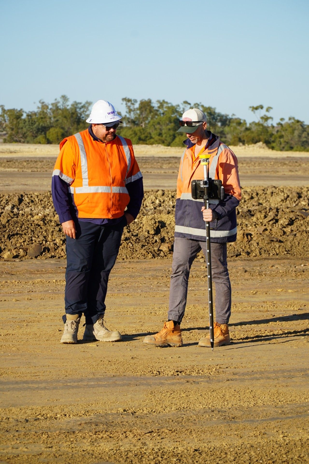 Two workers in orange safety vests and hard hats standing on a dirt worksite outdoors.