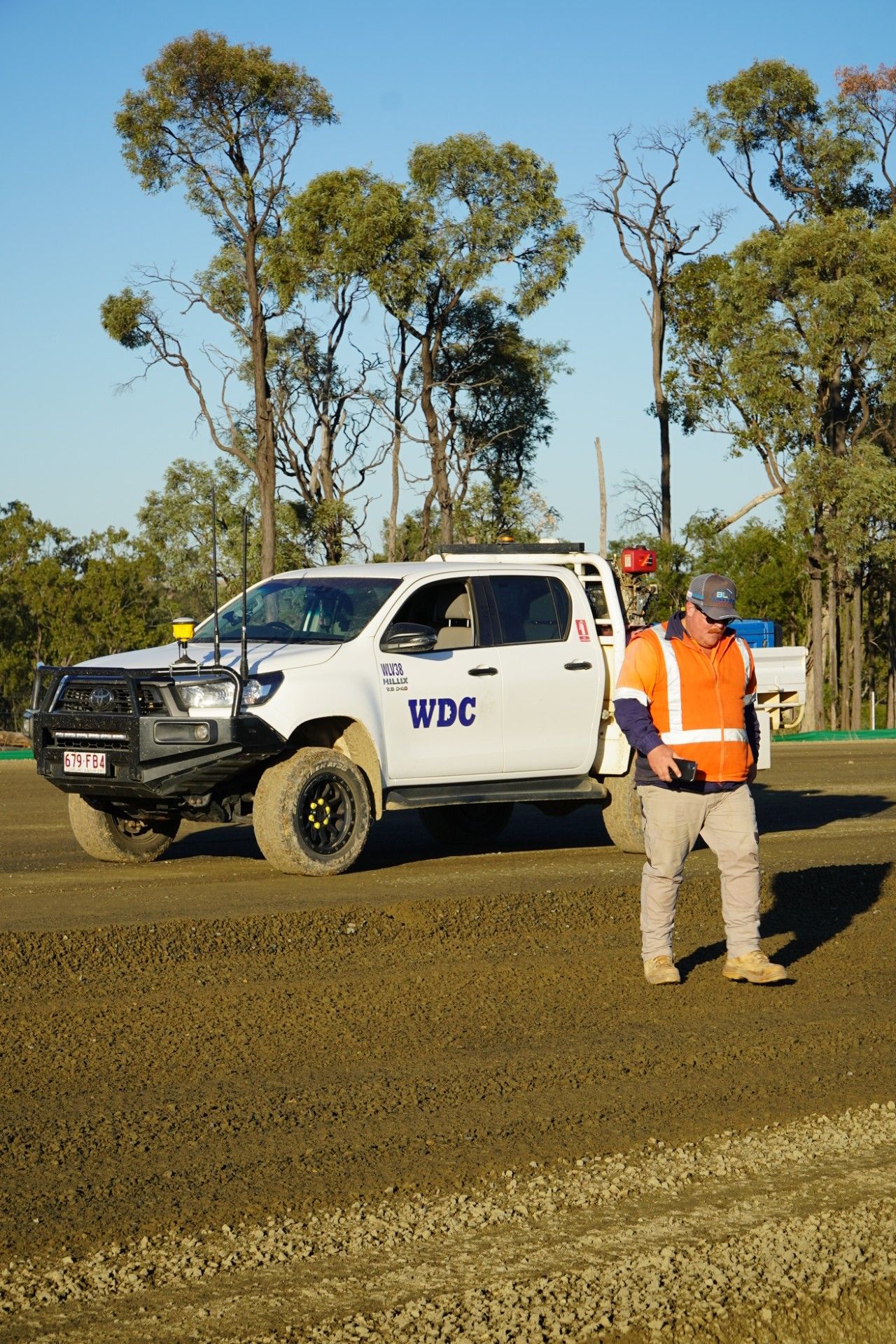 Worker in orange vest stands beside a white UDC pickup on a dirt road with trees in the background