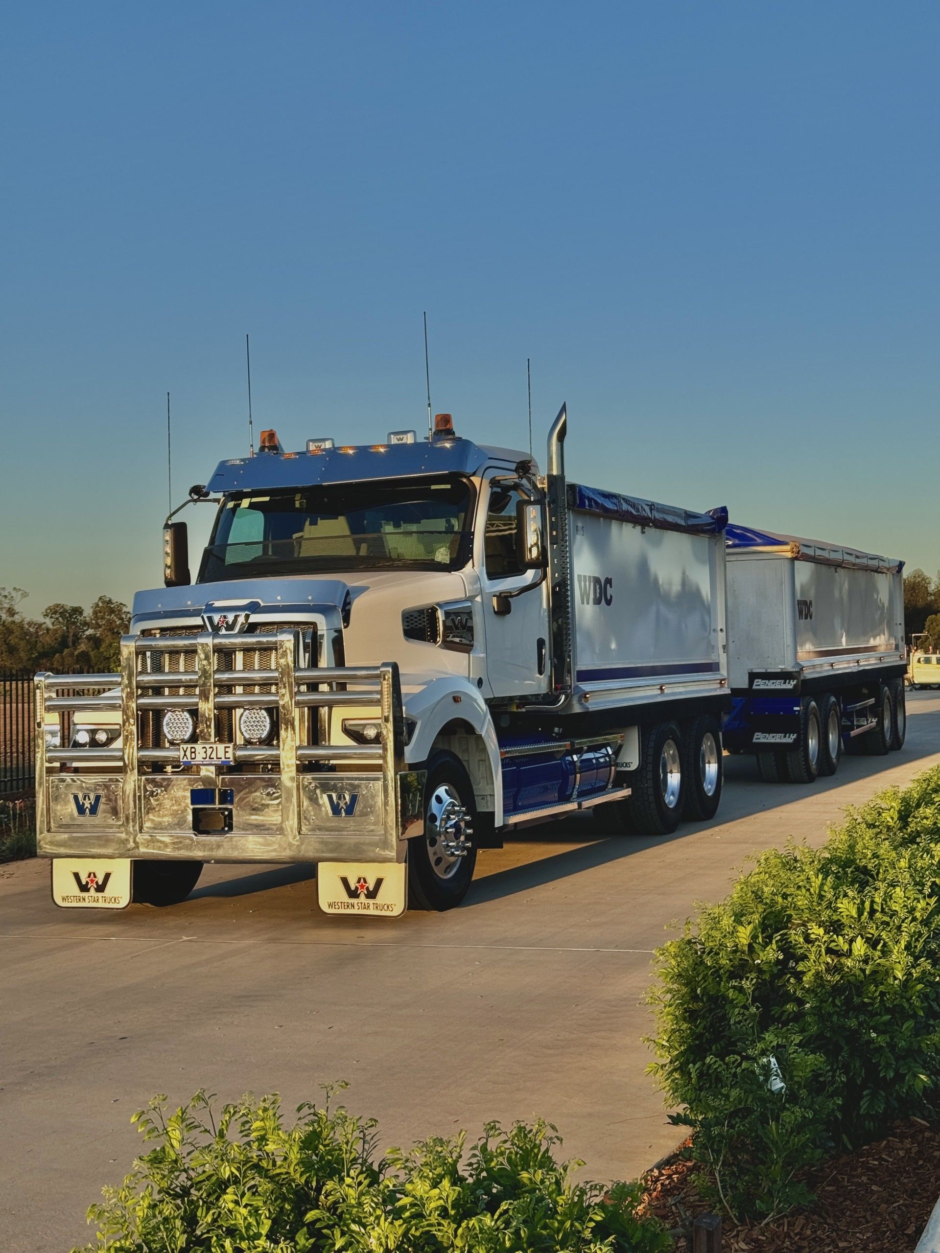 White dump truck with chrome bull bar parked on a road in bright sunlight, carrying a load in trailers