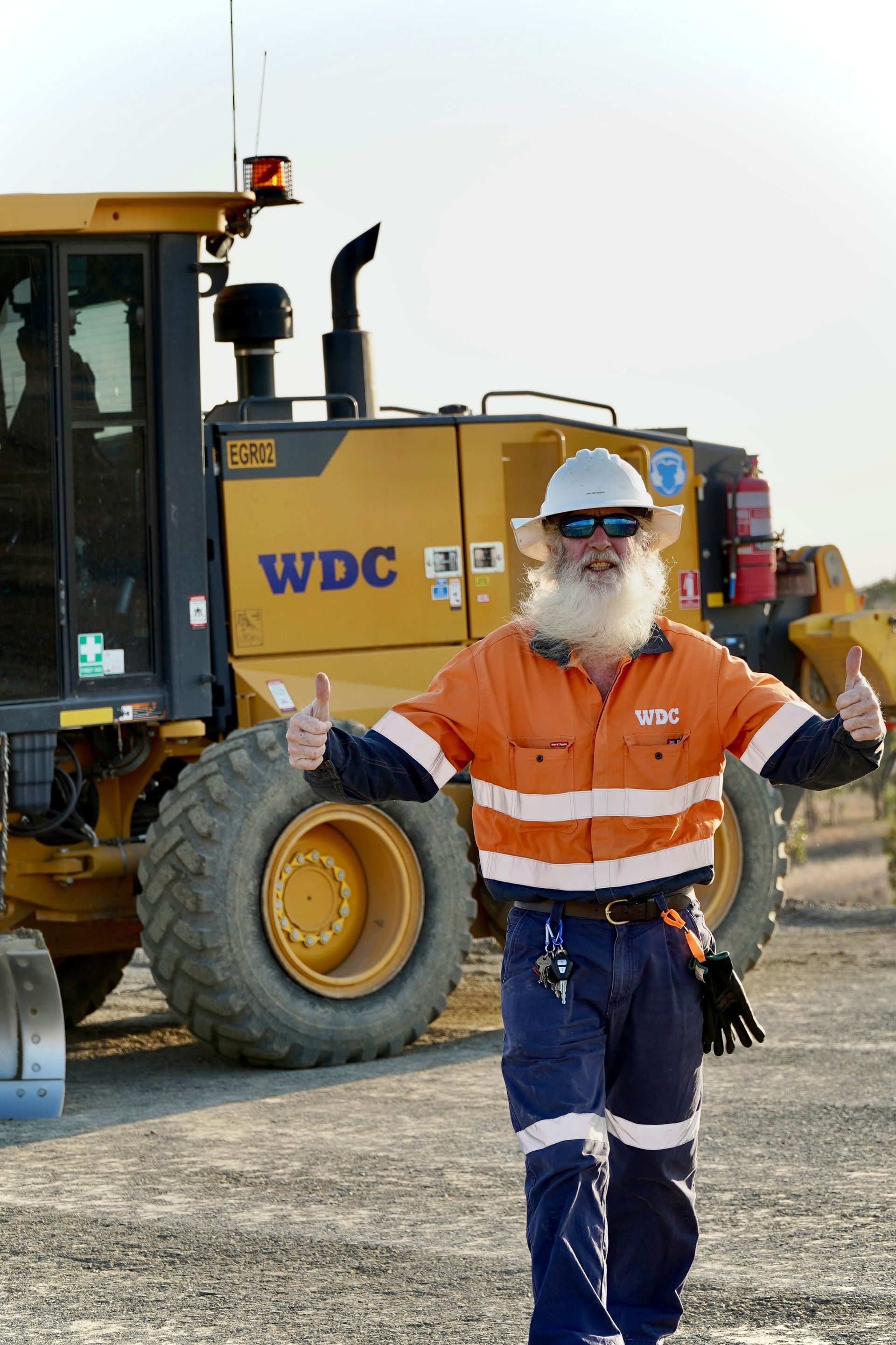 Construction worker in orange safety gear stands in front of a large loader, giving a thumbs-up.