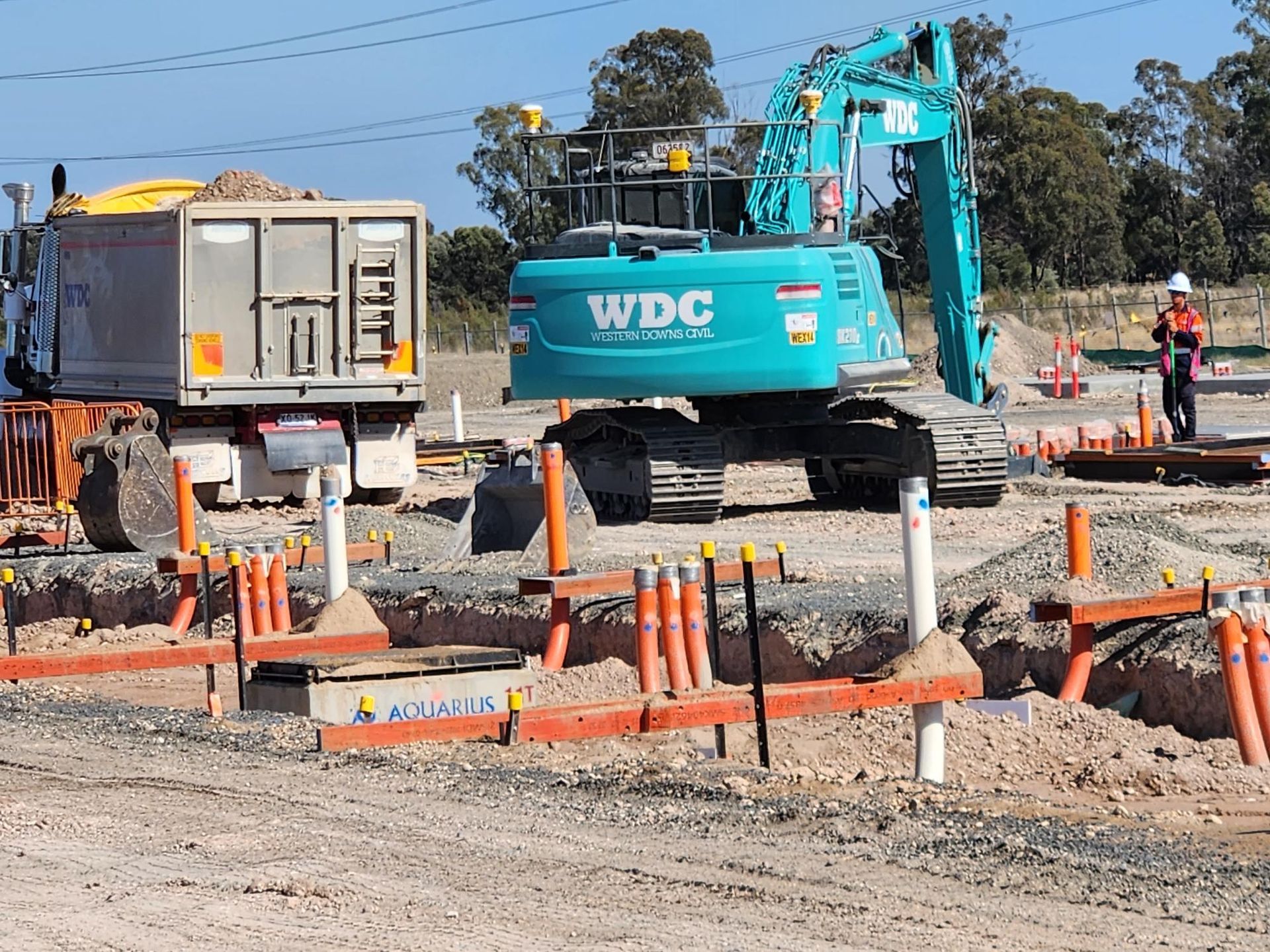 Construction site with excavator, truck, and orange safety barriers around a dug-up road.