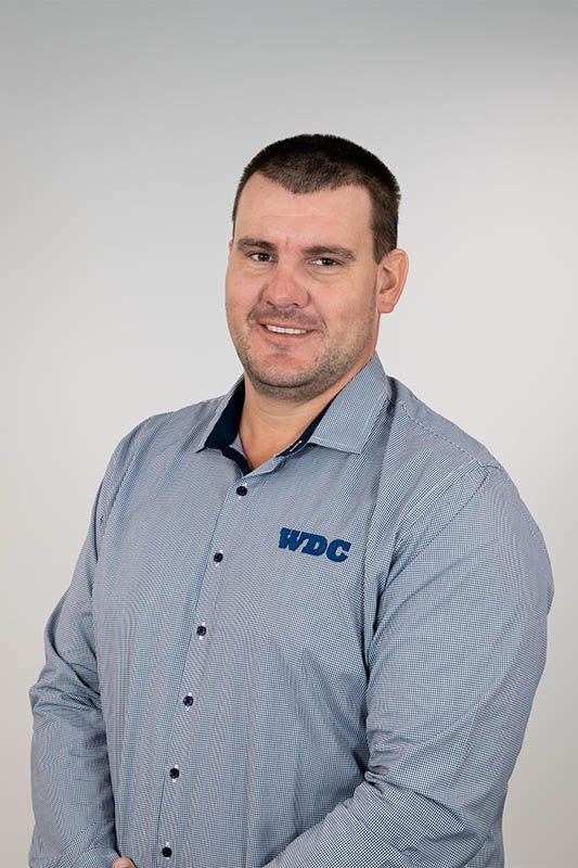 Smiling man in a blue button-up shirt with a logo, standing against a plain gray background