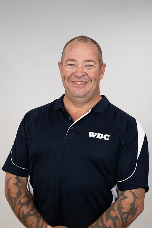 Man in navy WDC polo shirt standing against a light gray background, smiling with tattooed forearms visible