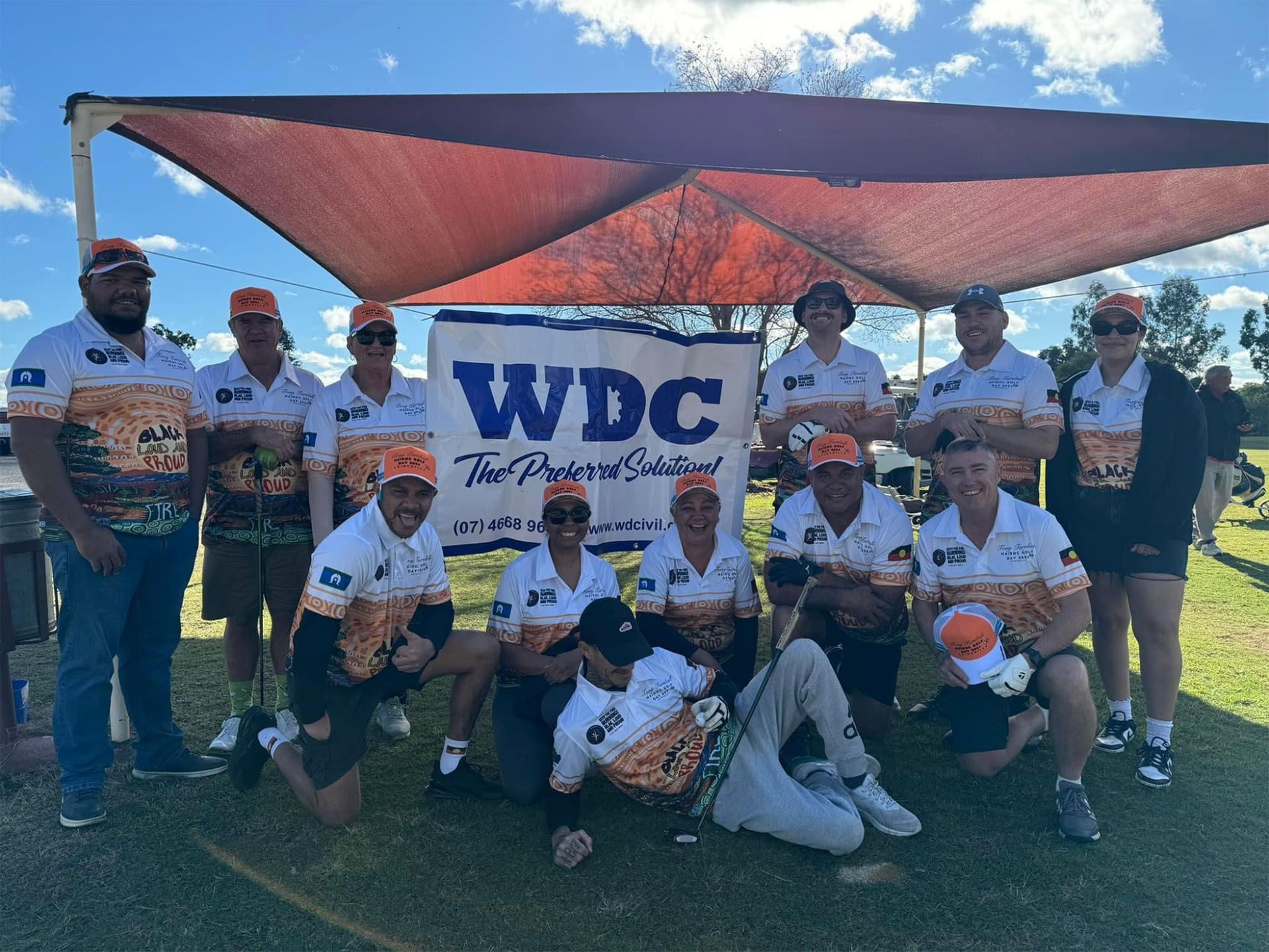 Group of people posing outdoors under a red shade canopy with a WDC banner at center