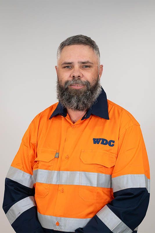 Man in orange high-visibility work jacket with navy sleeves, standing against a plain gray background.