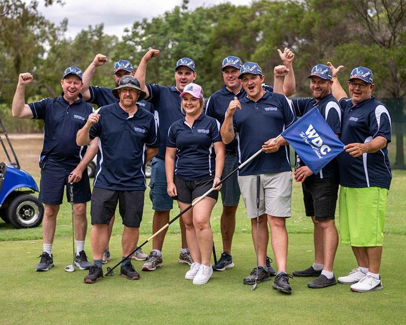 Golf team posing on a course, smiling and raising fists, one holding a blue flag.
