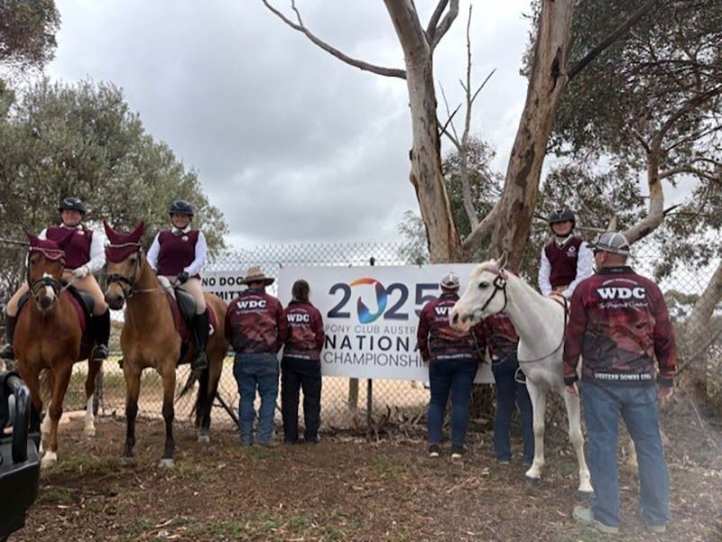 People on horseback and standing beside a banner at an outdoor equestrian event, with trees and cloudy sky.
