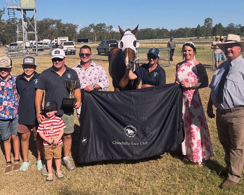 Group posing with a horse holding a black blanket at a racetrack, with a trophy in front.