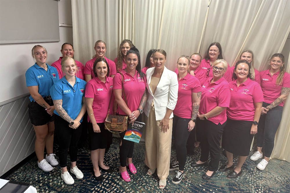 Group of smiling people in pink and blue shirts posing indoors, with two holding a plaque and gift bag.
